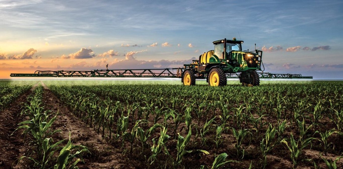 Pršilnik John Deere pri delu na koruznem polju s sončnim zahodom v ozadju