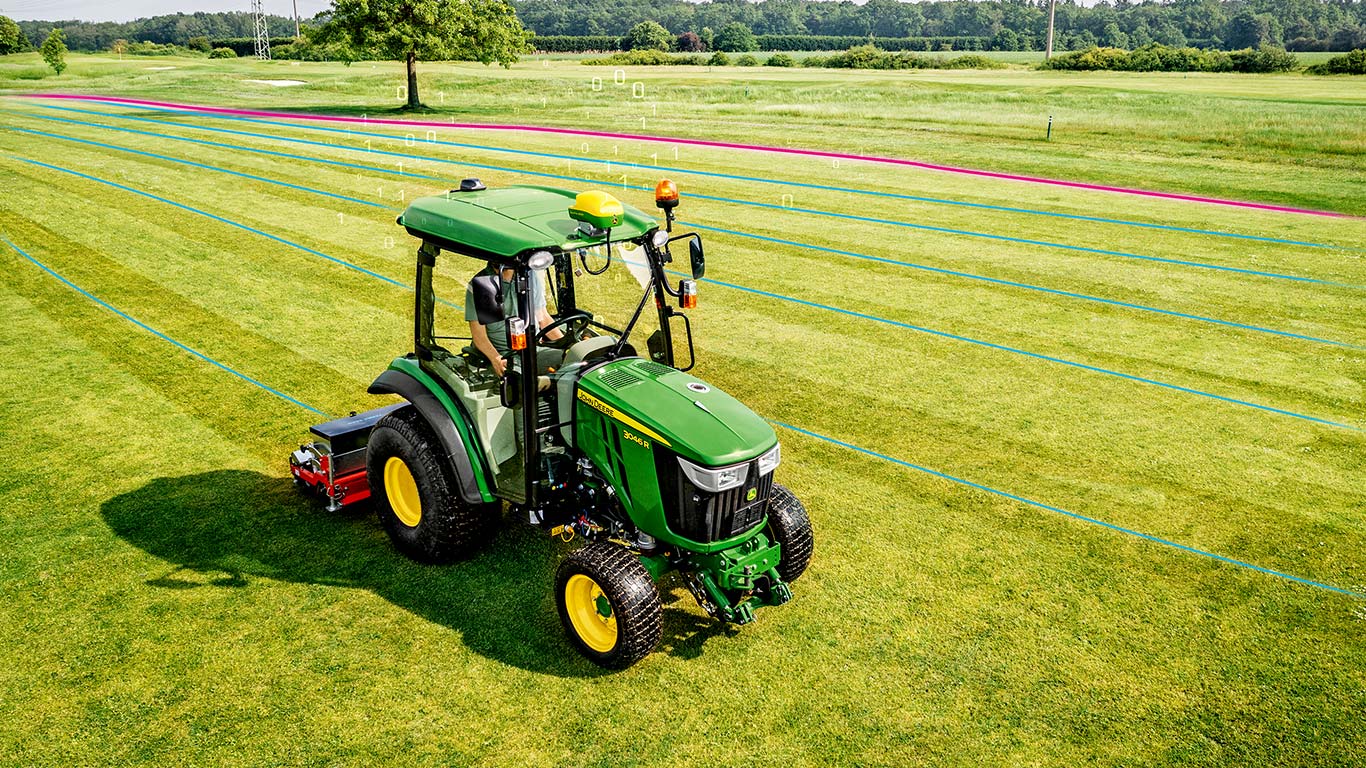 Vzdrževalec zelenic dela na travnati površini s kompaktnim večnamenskim traktorjem John Deere.