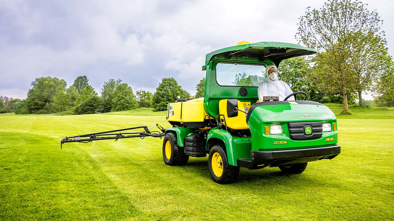 Vzdrževalec zelenic v zaščitnih oblačilih dela na zelenici igrišča za golf s škropilnikom John Deere.