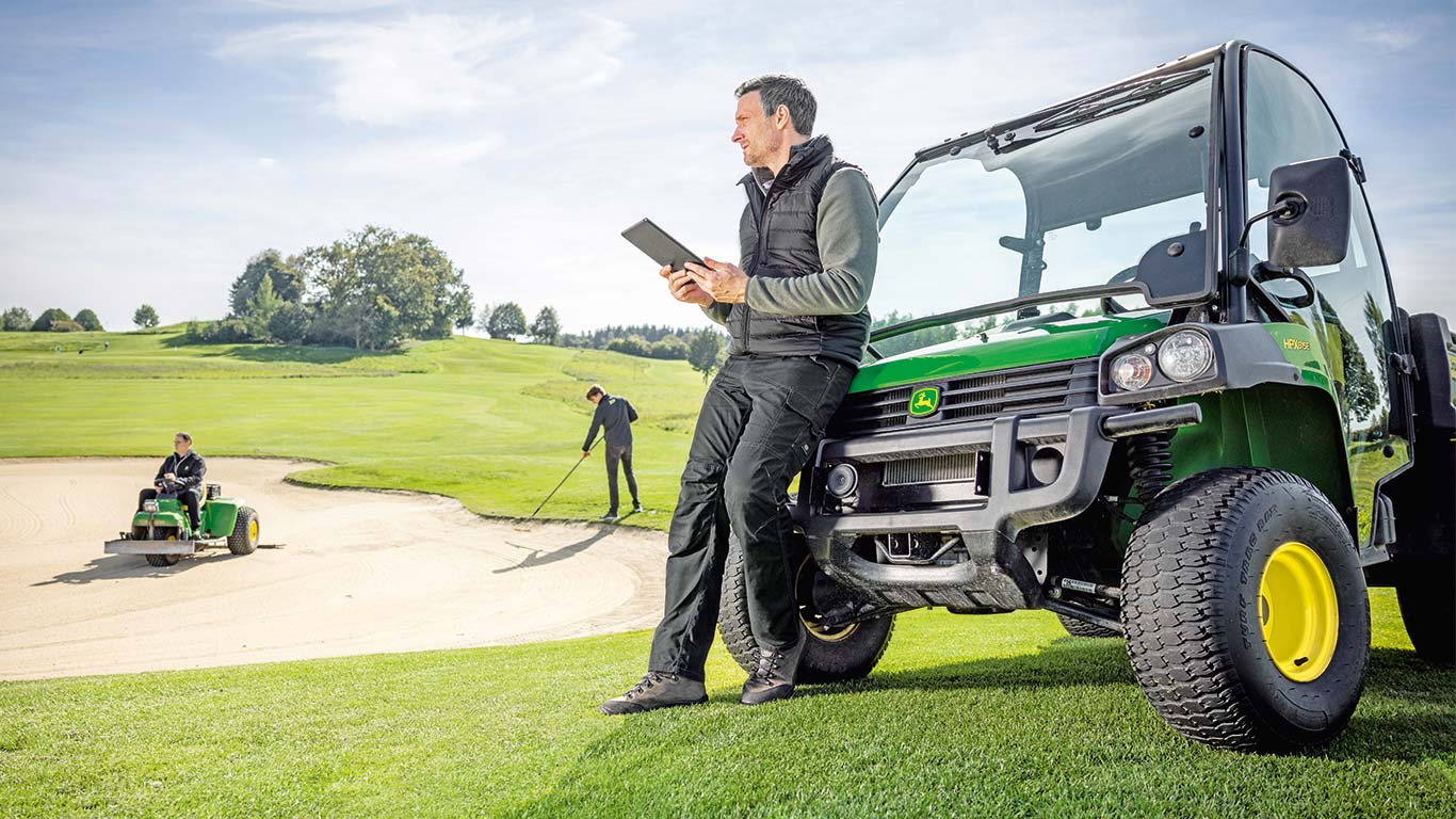 Vodja vzdrževanja zelenic preverja podatke na svoji tablici, medtem ko dva vzdrževalca zelenic delata na igrišču za golf v ozadju.