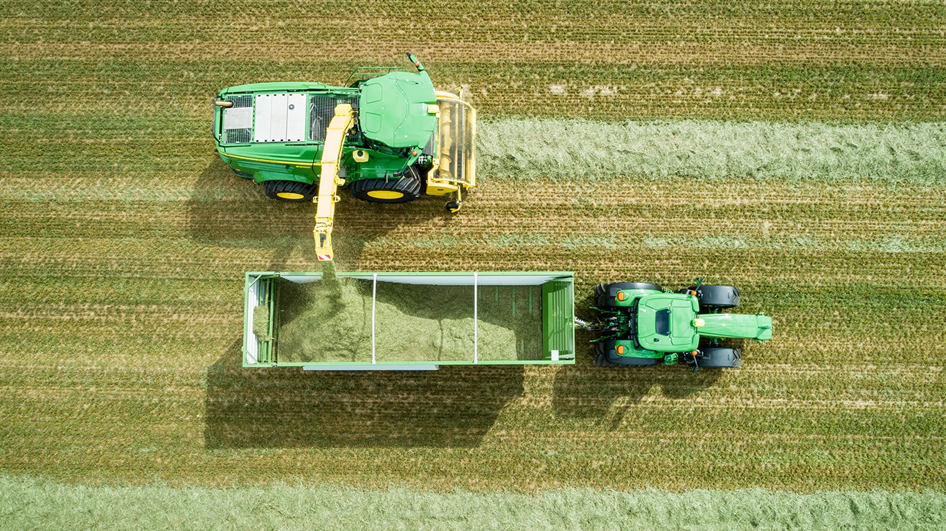 Fotografija iz zraka kombajna John Deere serije 8000 s samodejnim pogonom in strojem za pobiranje trave, ki jo silira v prikolico, ki jo vleče traktor John Deere