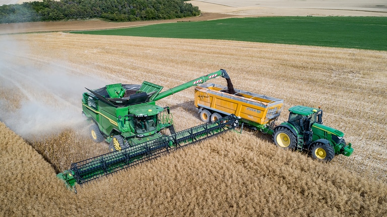 Kombajn John Deere serije X raztovarja repico v prikolico za seme, ki jo vleče traktor John Deere