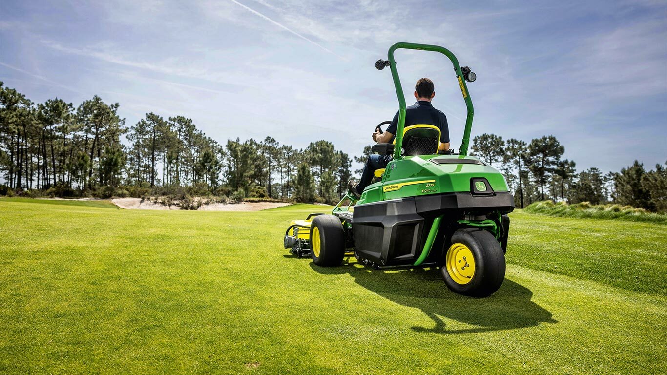 Oseba vozi zeleno električno kosilnico za zelenice John Deere 2775 E-Cut™ po dobro vzdrževanem igrišču za golf z drevesi in delno oblačnim nebom.