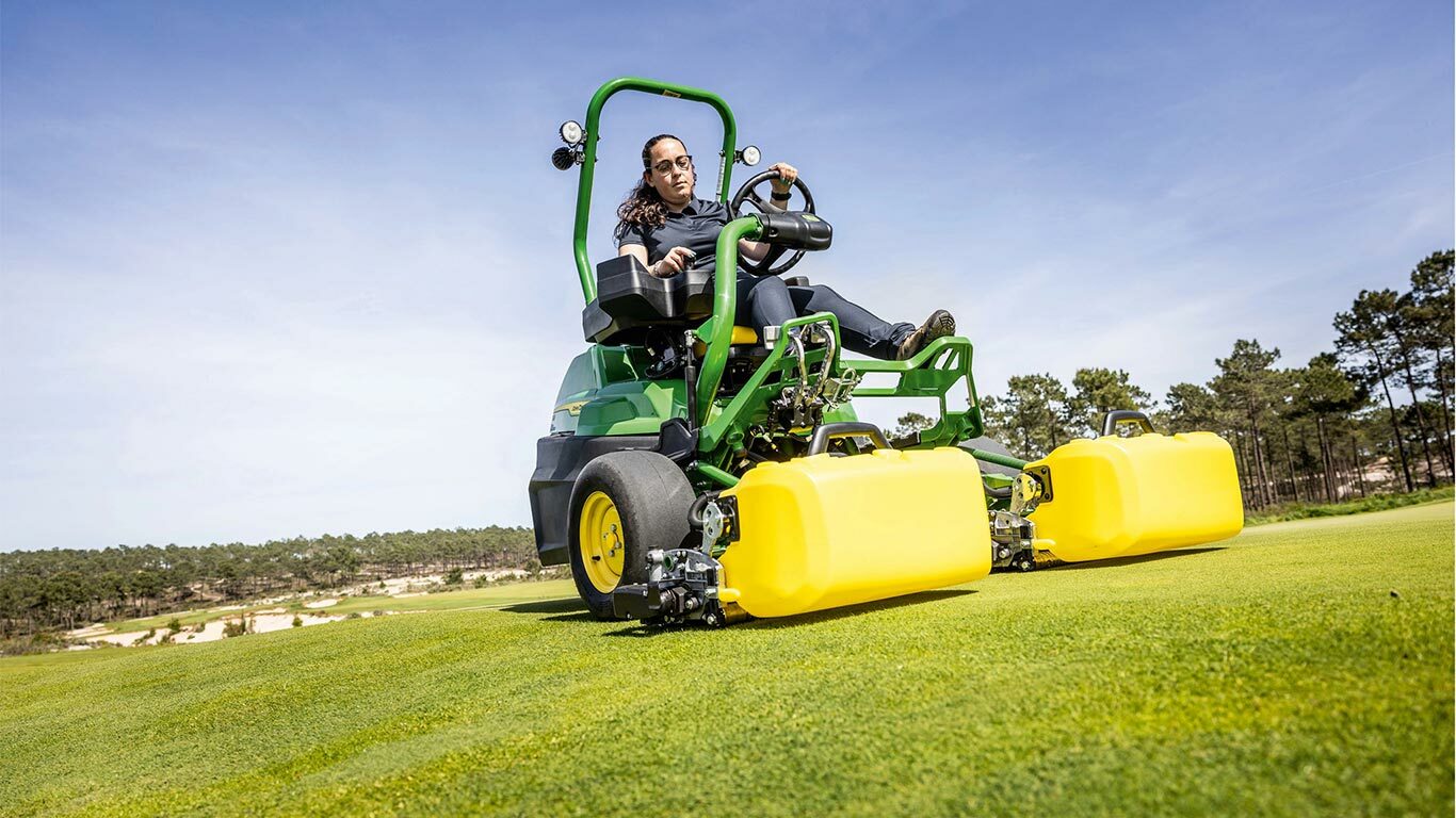 Oseba upravlja zeleno in rumeno električno kosilnico John Deere 2775 E-Cut™ s sprednjimi rezilnimi enotami, prekritimi z rumenimi ohišji na golf zelenici, z drevesi in jasnim modrim nebom v ozadju.