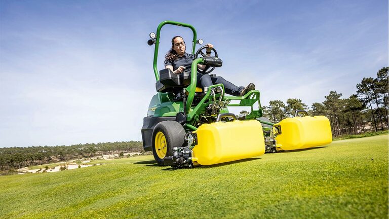 Oseba upravlja zeleno in rumeno električno kosilnico John Deere 2775 E-Cut™ s sprednjimi rezilnimi enotami, prekritimi z rumenimi ohišji na golf zelenici, z drevesi in jasnim modrim nebom v ozadju.