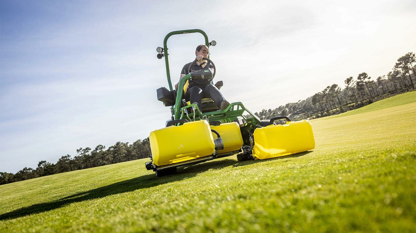Oseba upravlja zeleno in rumeno električno kosilnico John Deere 2775 E-Cut™ s tremi velikimi sprednjimi rezilnimi enotami, prekritimi z rumenimi ohišji na golf zelenici, z drevesi in delno oblačnim nebom v ozadju.