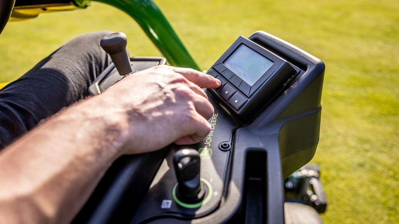 Bližnji posnetek dlani na nadzorni plošči TechControl električne kosilnice John Deere 2775 E-Cut™, ki pritiska gumbe na digitalnem prikazovalniku s krmilno palico in drugimi vidnimi upravljalnimi elementi.