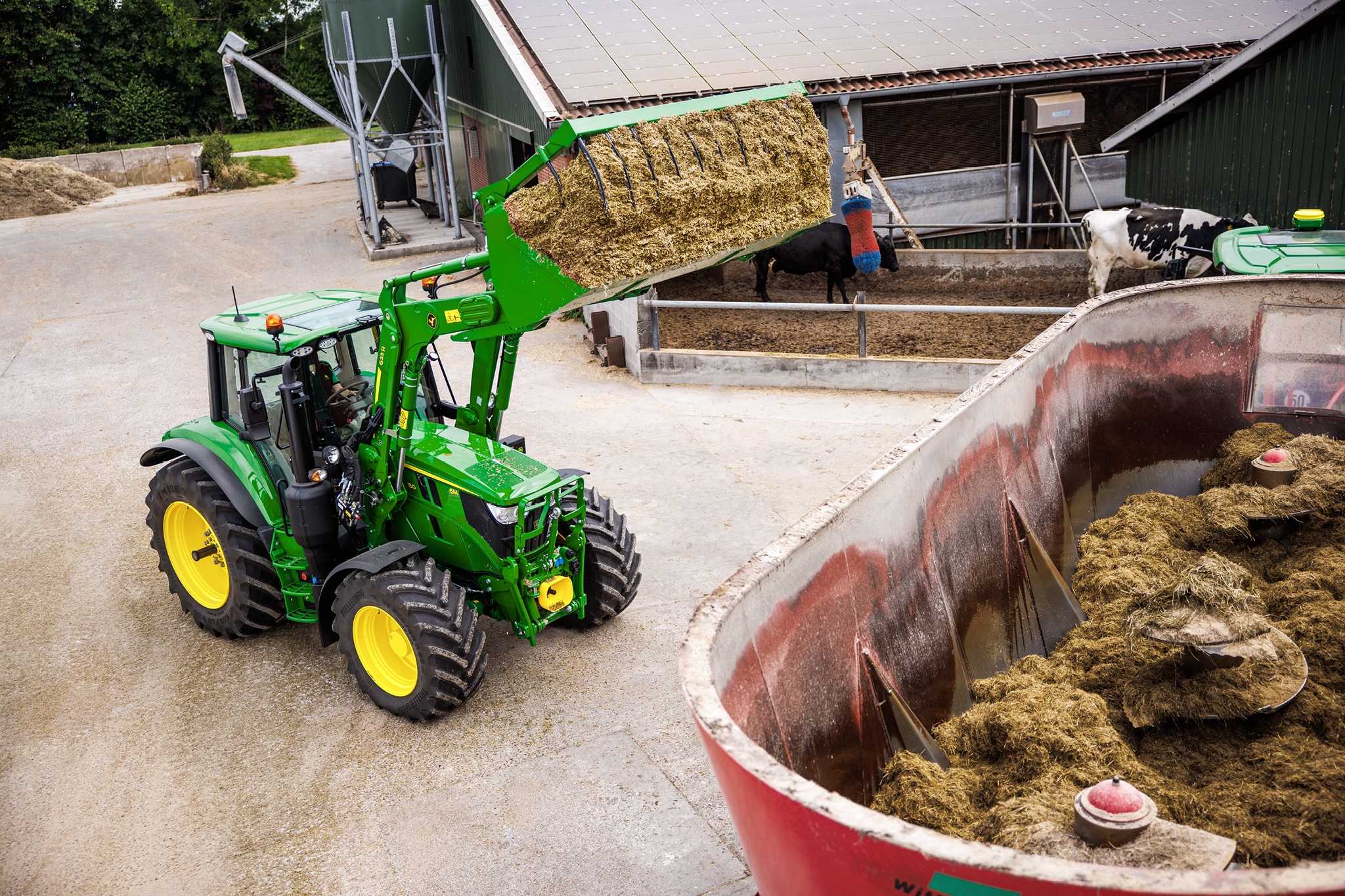Traktor John Deere serije 6M s sprednjim nakladalnikom natovarja silažo v prikolico mešalnika na dvorišču kmetije.