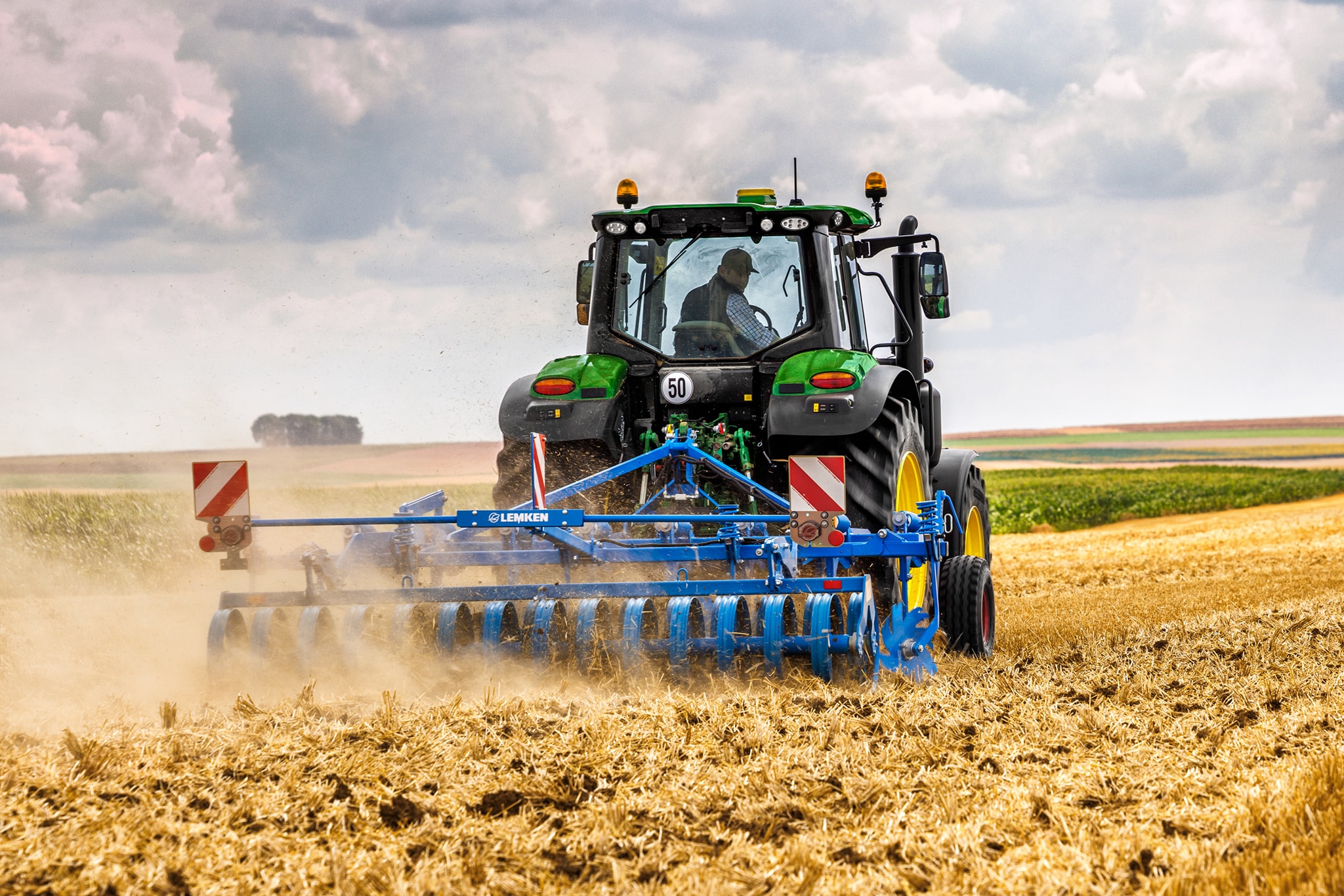 Traktor John Deere serije 6M obdeluje požeto polje s kolutno brano, pogled od zadaj na prašnem polju.