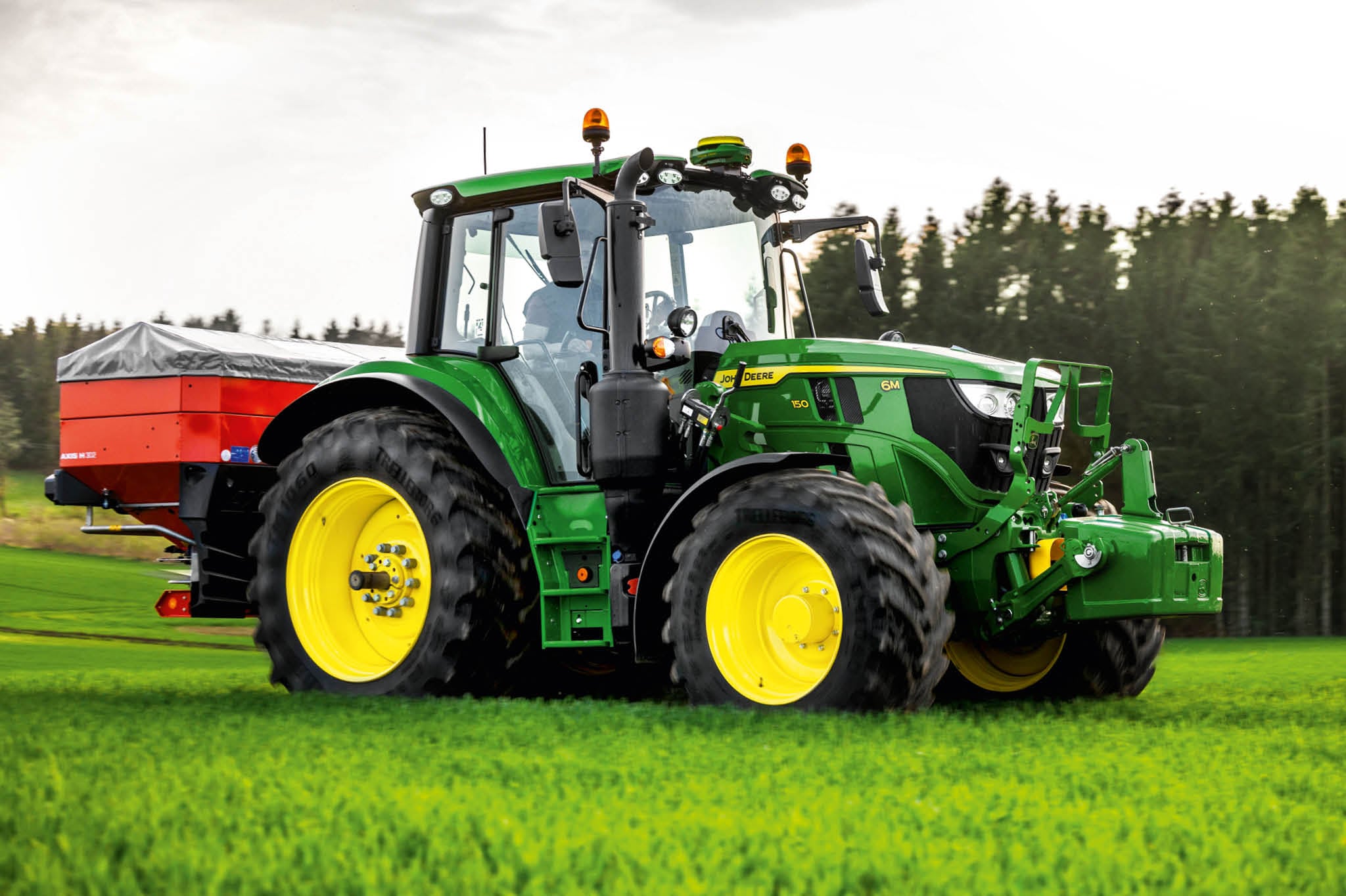 Traktor John Deere serije 6M z zadnjim trosilnikom, ki deluje na travi blizu roba gozda.