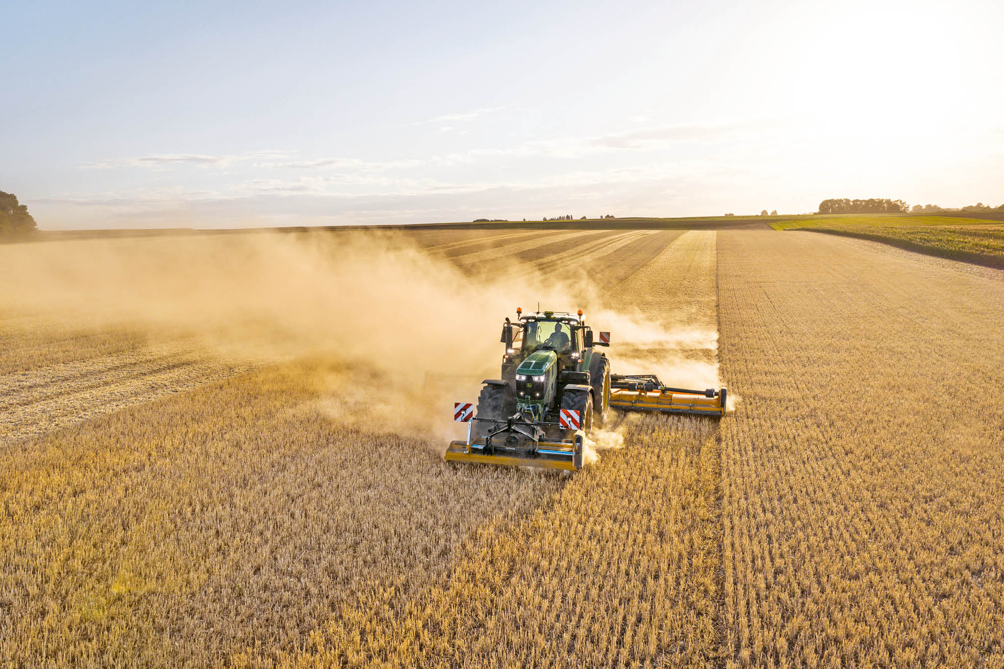 Traktor John Deere serije 6M vleče cisterno za gnojevko po podeželski cesti, obdani s travnikom in poljem.