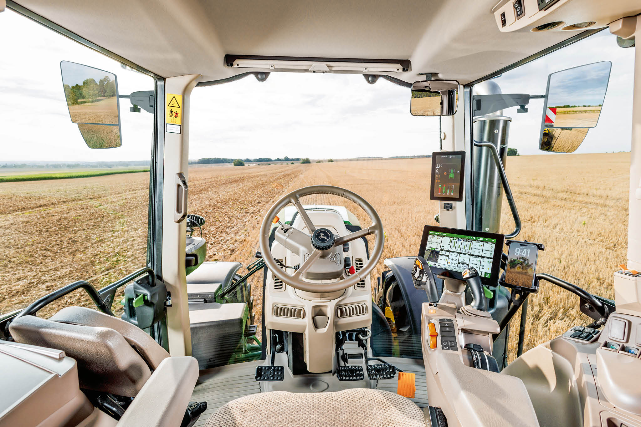 Prikaz notranjosti kabine traktorja John Deere serije 6M, ki prikazuje volan, upravljalne prikazovalnike in strnišče pred traktorjem.