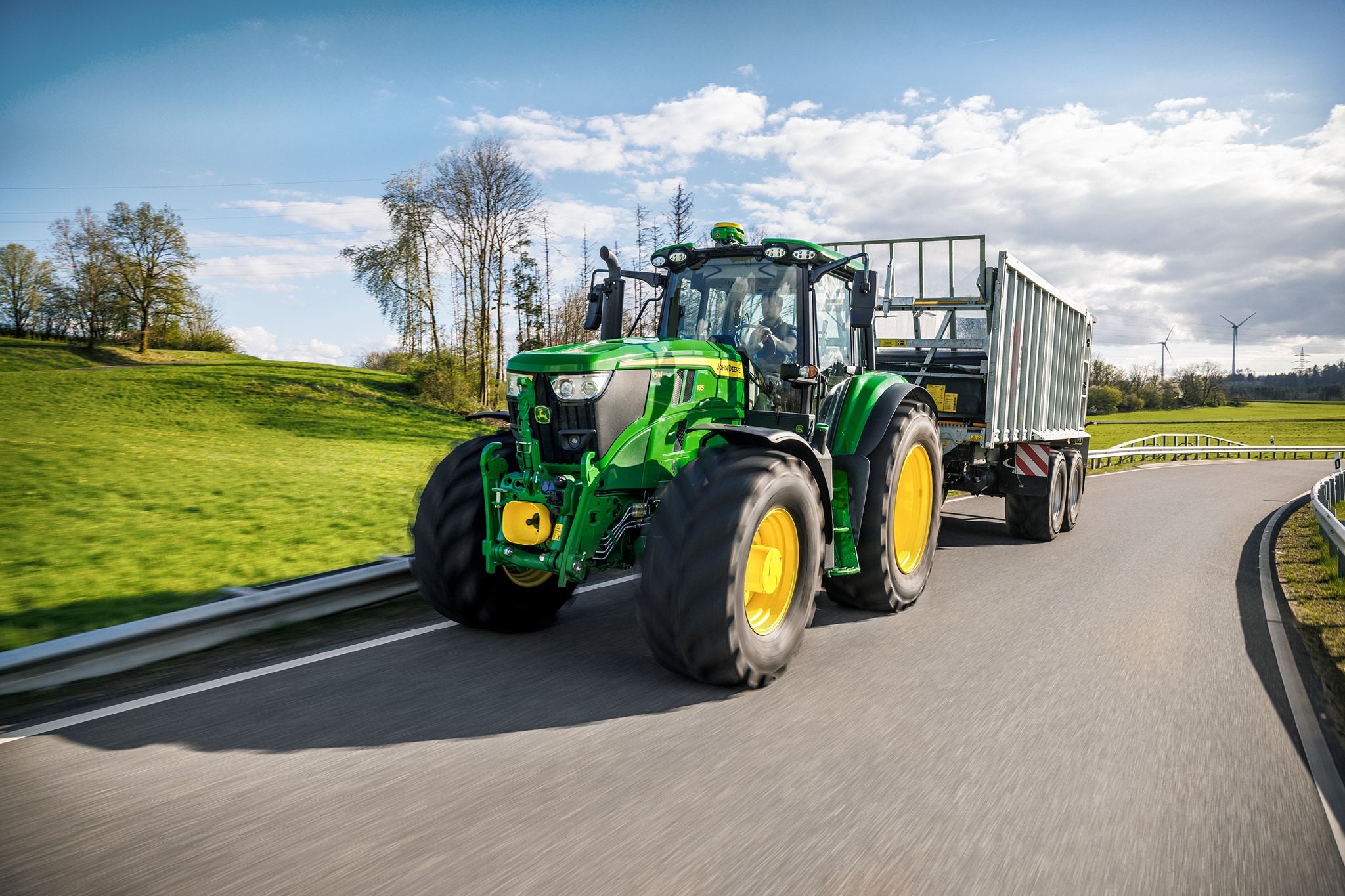 Traktor John Deere serije 6M prevaža prikolico z visokimi stranicami po asfaltirani podeželski cesti.