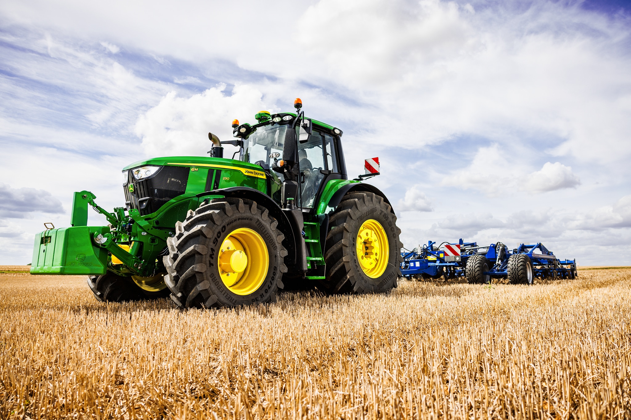 Traktor John Deere serije 6M vleče težak priključek za oranje po strnišču pod oblačnim nebom.