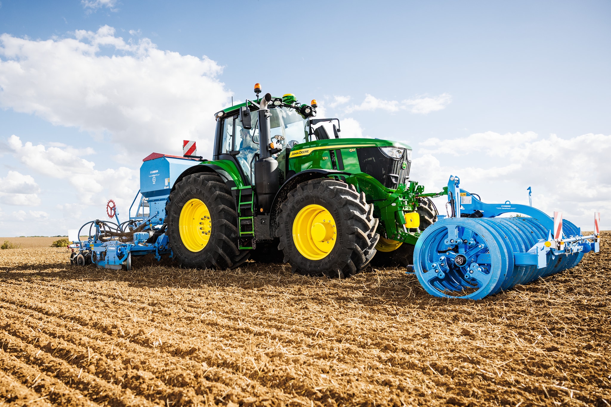 Traktor John Deere serije 6M obdeluje polje s priključenim velikim modrim valjastim priključkom.