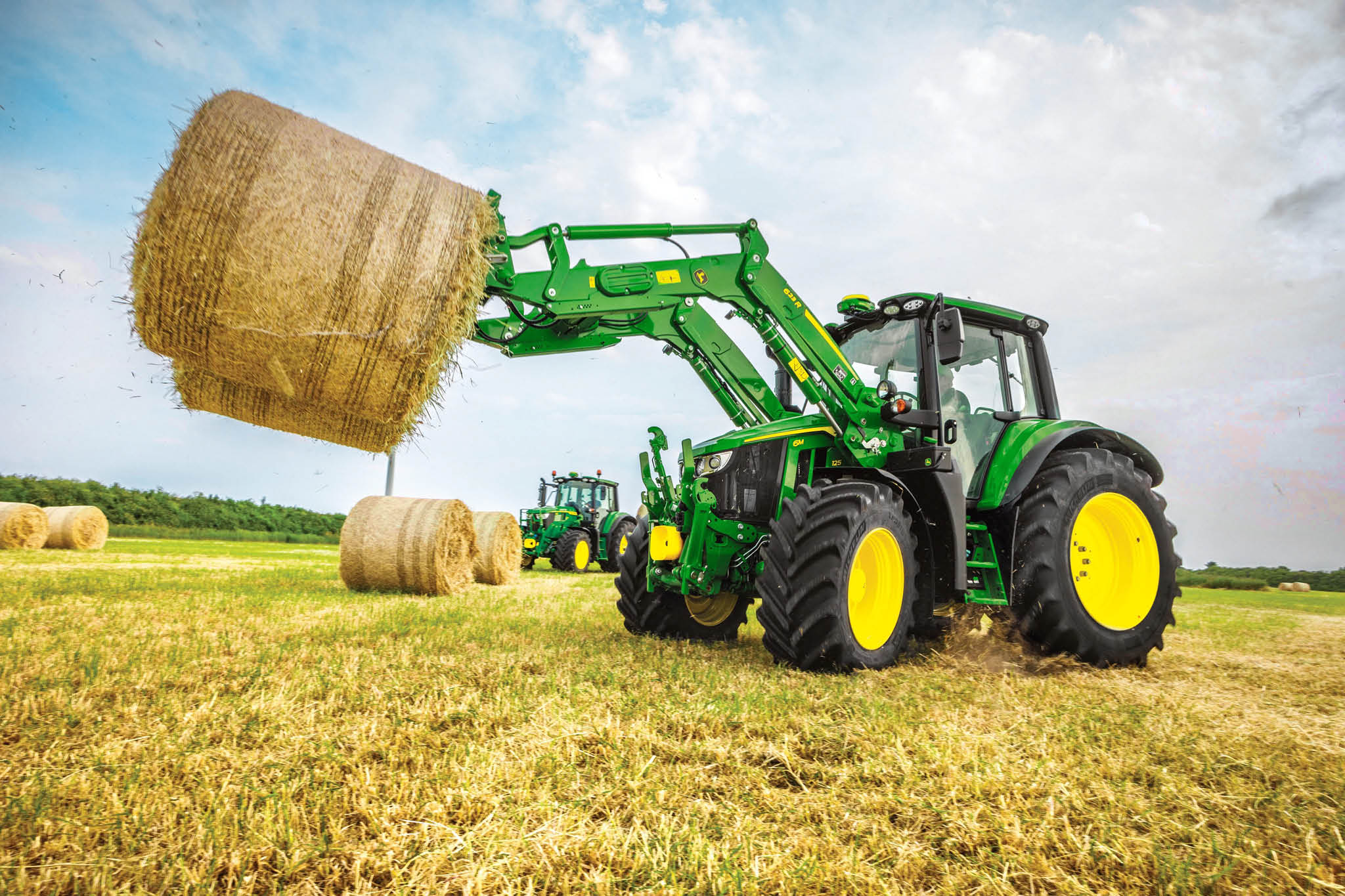 Traktor John Deere serije 6M dviguje veliko okroglo balo s sprednjim nakladalnikom na požetem polju pod modrim nebom.