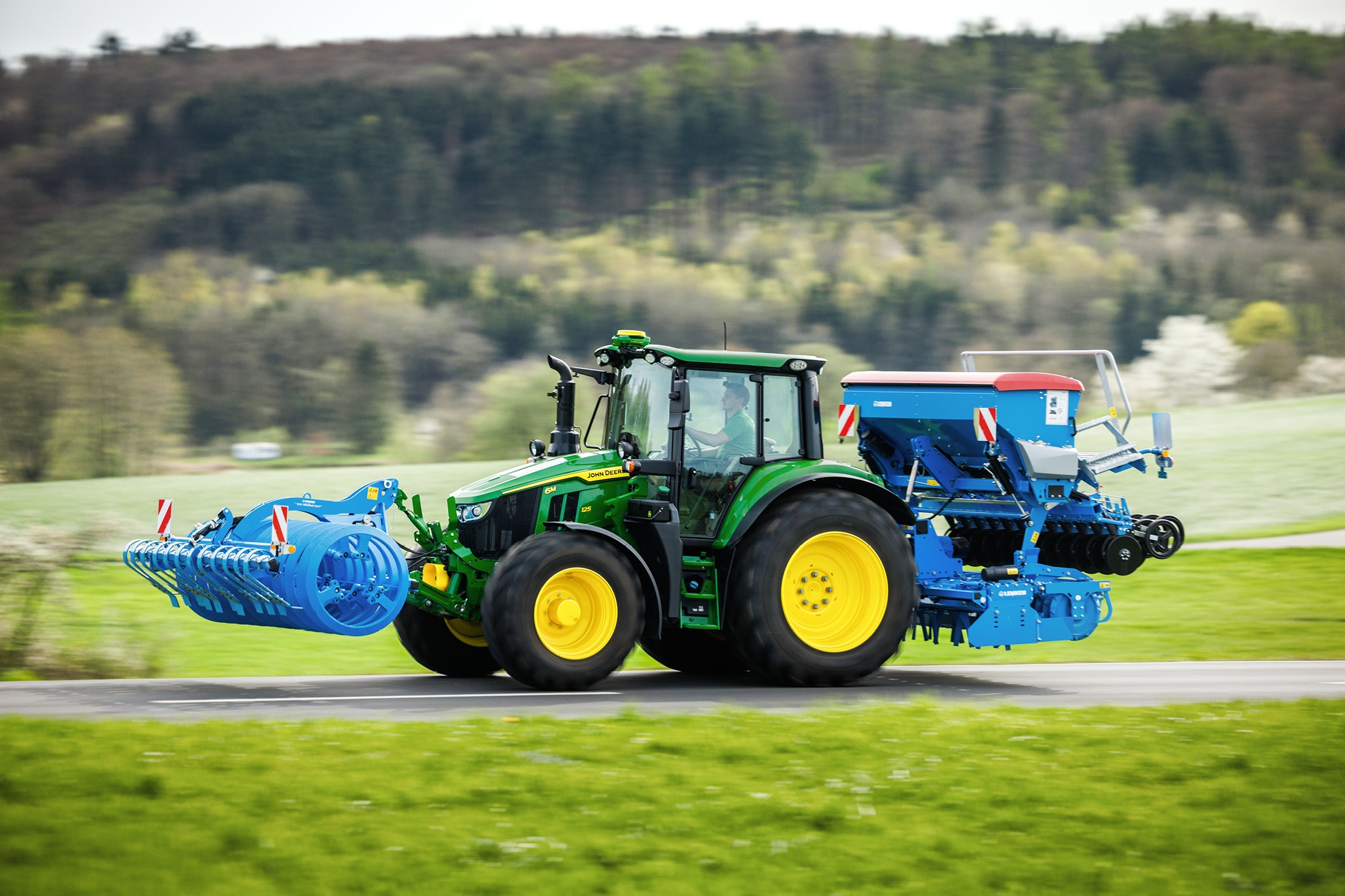 Traktor John Deere serije 6M prevaža spredaj nameščen priključek in zadnji sejalnik po podeželski cesti.