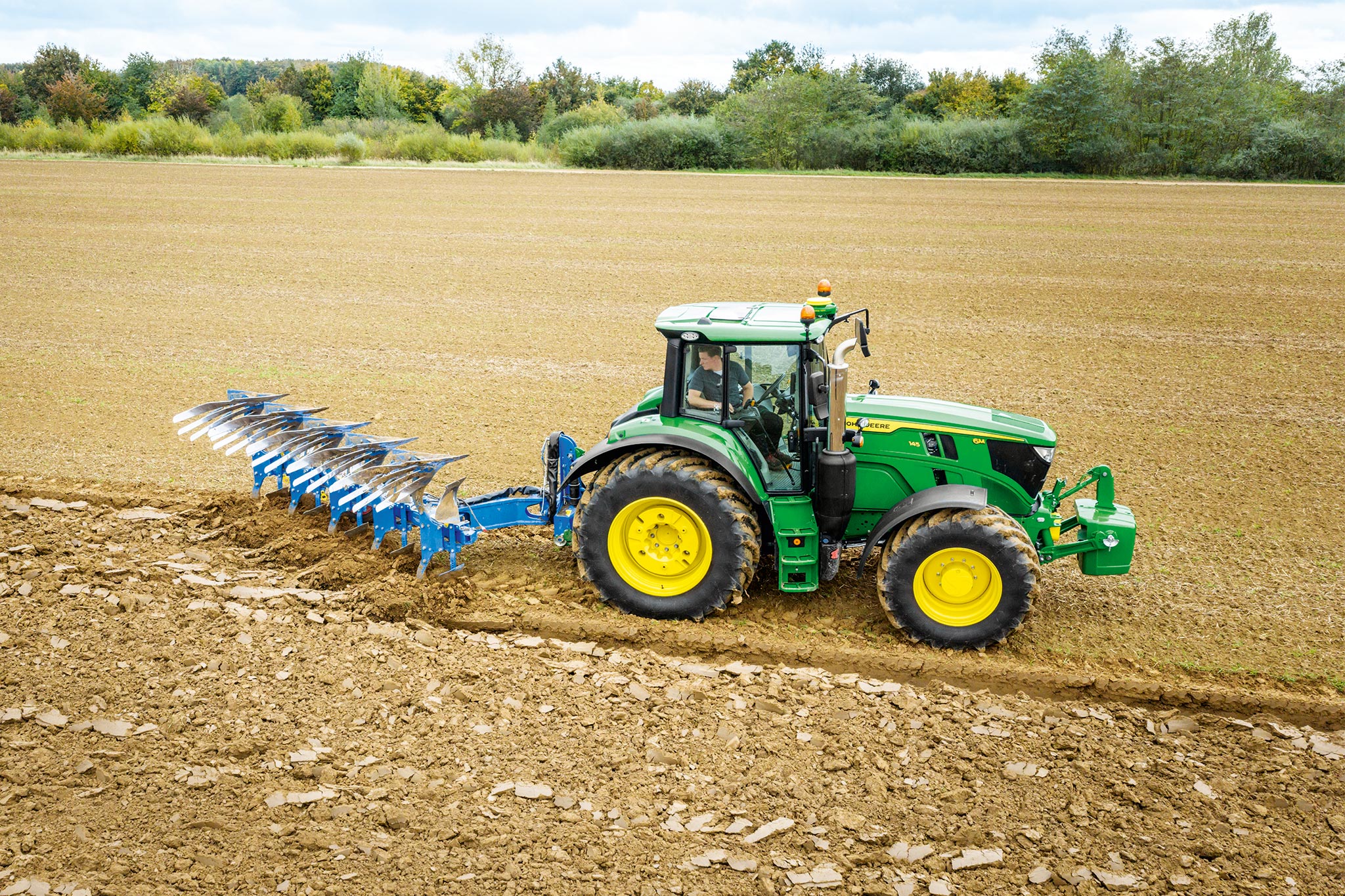 Traktor John Deere serije 6M obdeluje zorano polje z zadaj nameščenim orodjem za obdelavo tal pod oblačnim nebom.