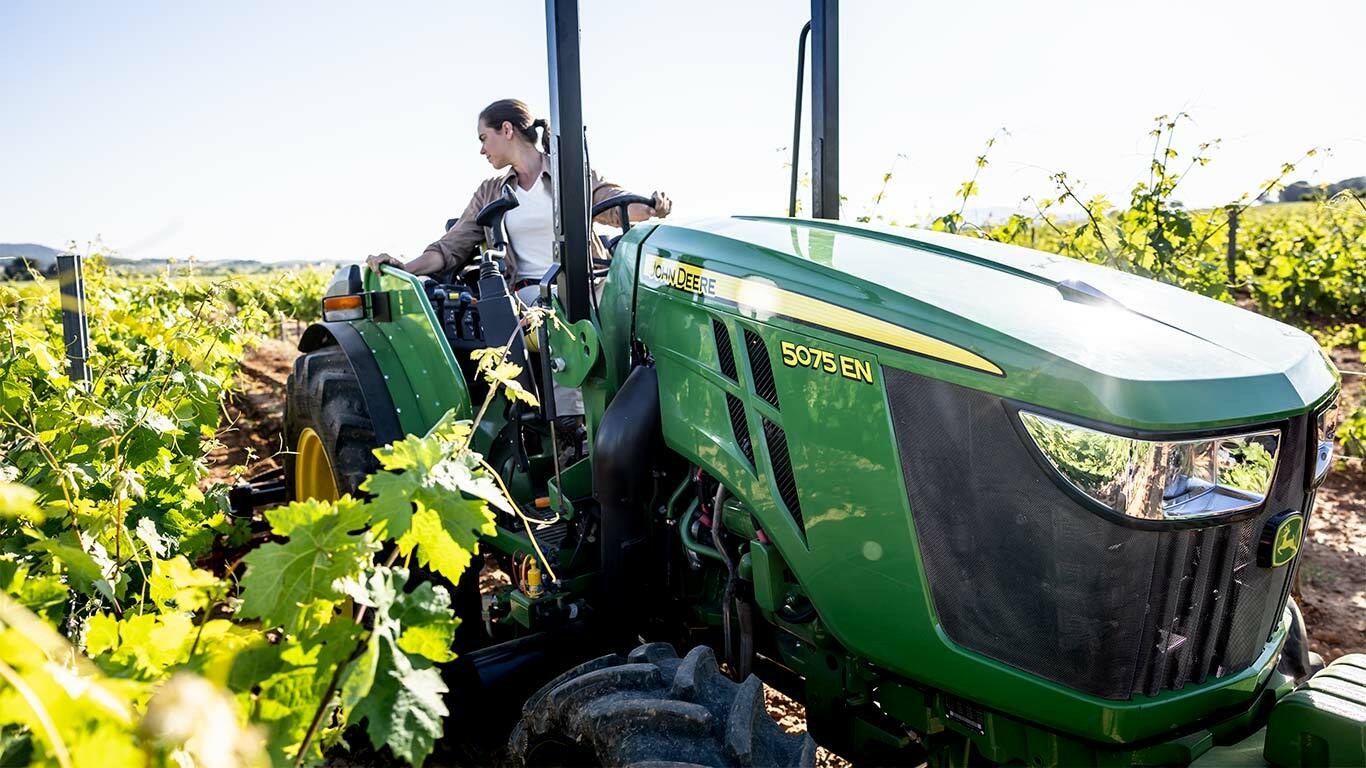 Traktor John Deere 5075EN v vinogradu, obdan z zelenimi trtami pod svetlim soncem.