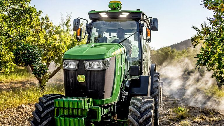 S soncem obsijan zeleno-rumeni traktor John Deere 5075EN vozi po poti skozi sadovnjak.