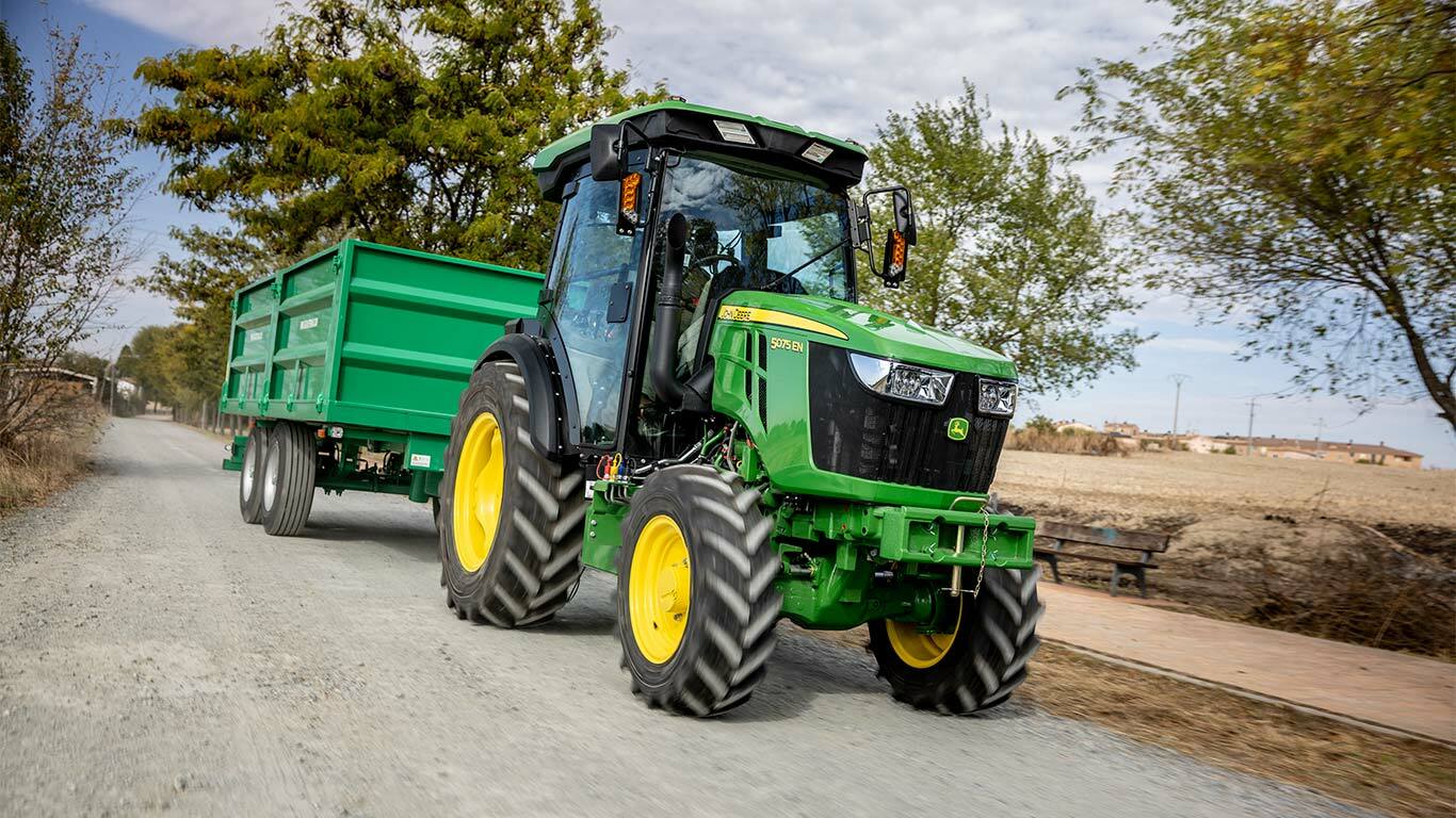 Traktor John Deere 5075EN vleče zeleno prikolico po podeželski makadamski cesti z drevesi v ozadju.