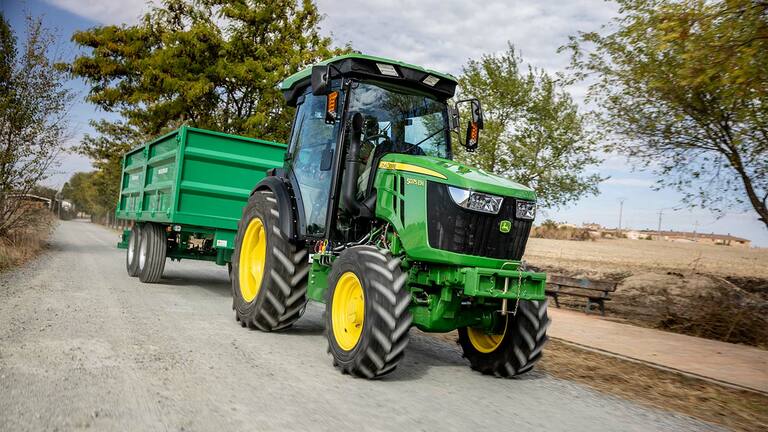 Traktor John Deere 5075EN vleče zeleno prikolico po podeželski makadamski cesti z drevesi v ozadju.