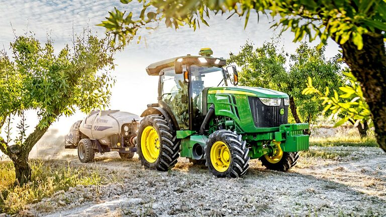 Zeleno-rumeni Traktor John Deere 5090EN na sončen dan vleče pršilnik skozi sadovnjak.