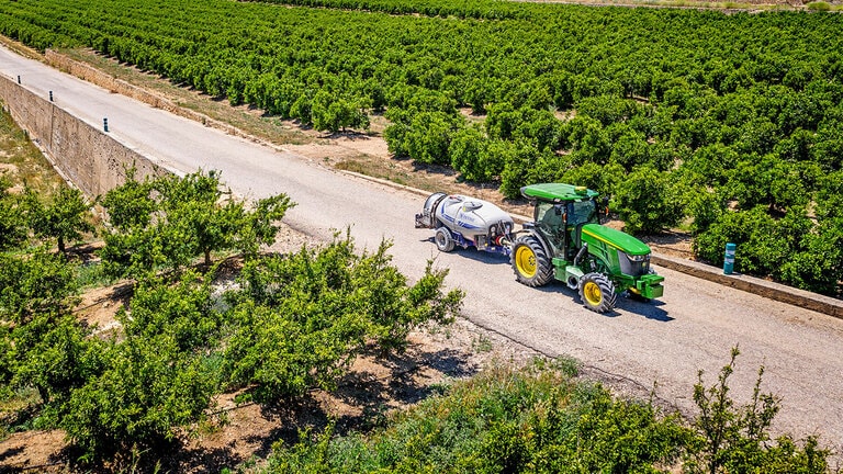 Zeleno-rumeni Traktor John Deere 5090EN na sončen dan vleče pršilnik po asfaltirani poti med vrstami v sadovnjaku.