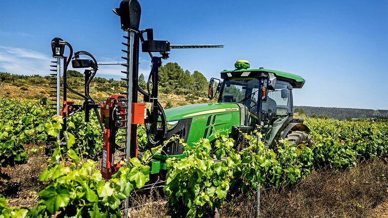 Zeleno-rumeni traktor John Deere 5090EN, opremljen z orodjem za obrezovanje, na sončen dan opravlja delo v vinogradu.