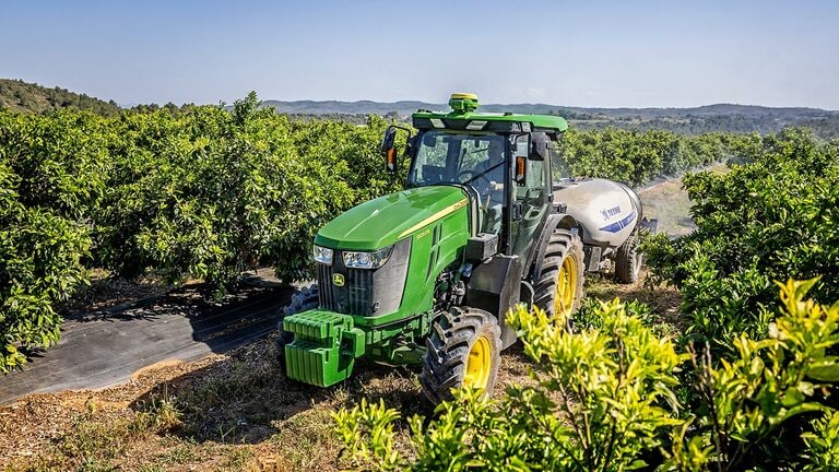 Zeleno-rumeni traktor John Deere 5105EN vleče pršilnik skozi sadovnjak z bujno rastjo, za njim se razprostirajo valoviti griči.
