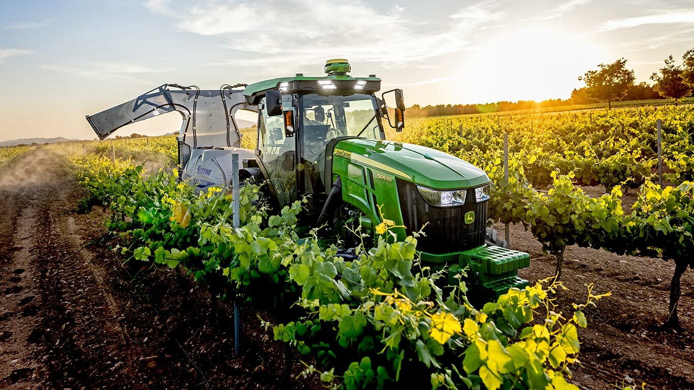 Zeleno-rumeni traktor John Deere 5105EN, ki dela v vinogradu ob sončnem zahodu in škropi poljščine med vrstami vinske trte.