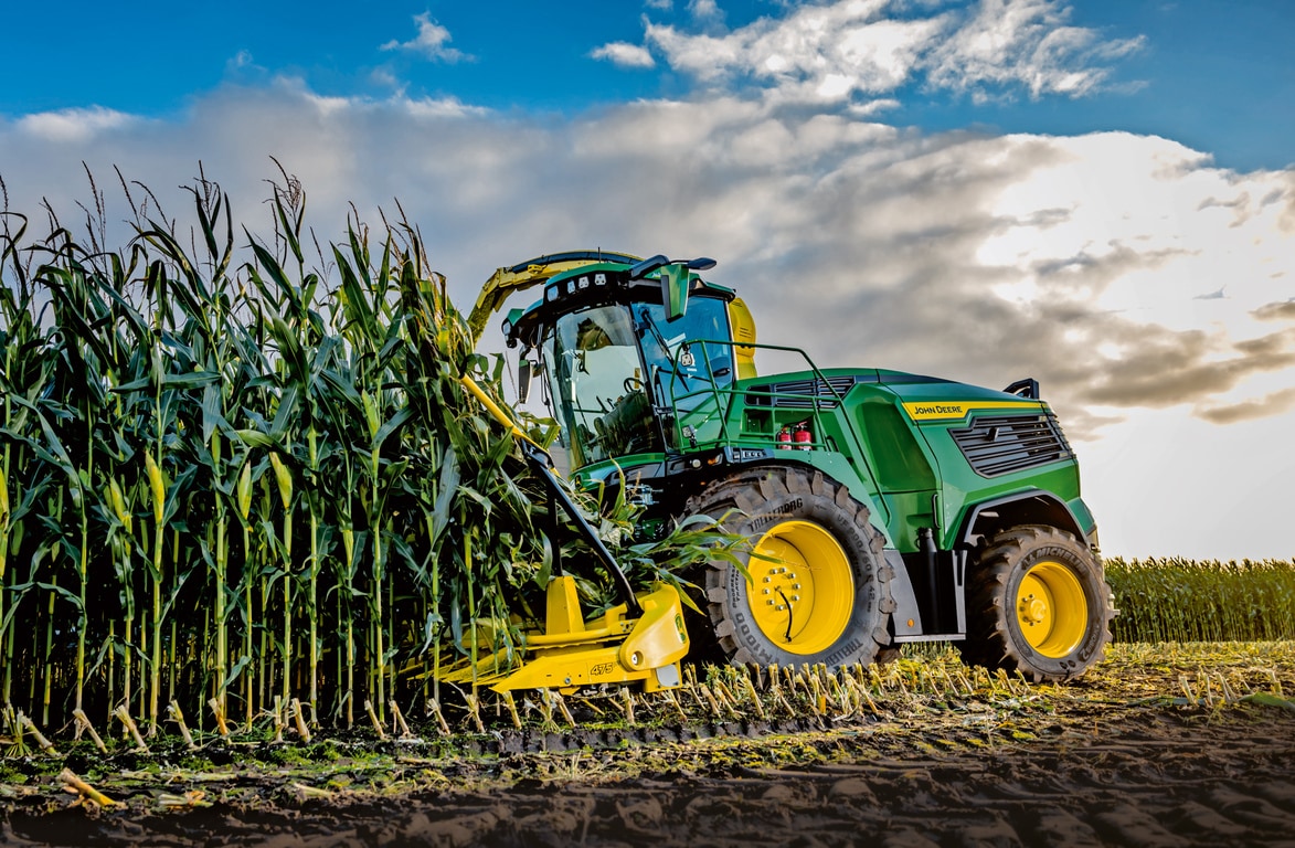 Silažni kombajn John Deere pri spravilu koruze na polju pod modrim nebom z oblaki.