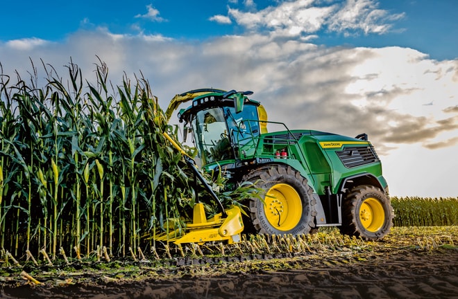 Silažni kombajn John Deere pri spravilu koruze na polju pod modrim nebom z oblaki.
