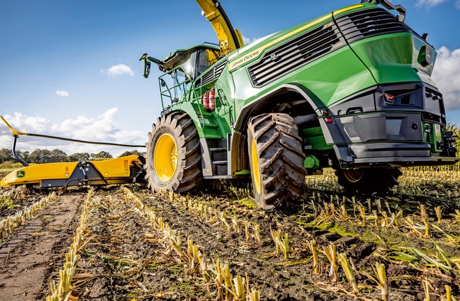 Silažni kombajn John Deere med delom na pospravljenem koruznem polju, pri čemer so na jasnem modrem nebu vidne velike pnevmatike in rezalni mehanizem.