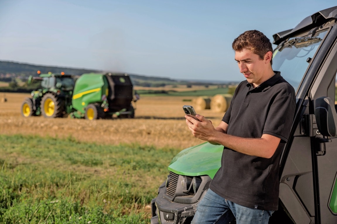 Oseba preverja podatke o dokumentaciji za bale na mobilnem telefonu, v ozadju pa je balirka John Deere VR