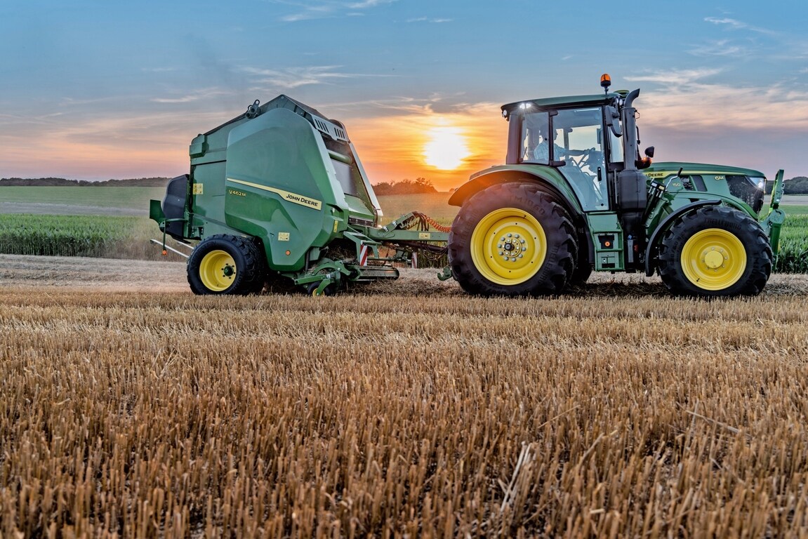 Traktor John Deere 6M 150 ob sončnem zahodu na polju slame vleče balirko John Deere V462M